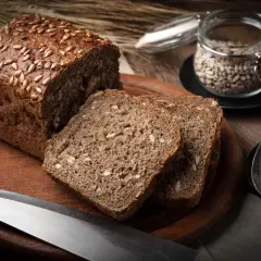 Bochenek chleba żytniego z ziarnami słonecznika, pokrojony na kromki, obok nasion i noża.