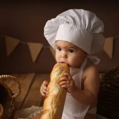 Niemowlę w czepku kucharskim gryzie bagietkę, siedząc obok koszyka z pieczywem.