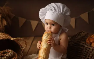 Niemowlę w czepku kucharskim gryzie bagietkę, siedząc obok koszyka z pieczywem.