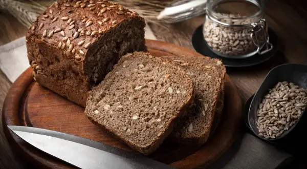 Bochenek chleba żytniego z ziarnami słonecznika, pokrojony na kromki, obok nasion i noża.