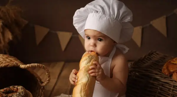 Niemowlę w czepku kucharskim gryzie bagietkę, siedząc obok koszyka z pieczywem.